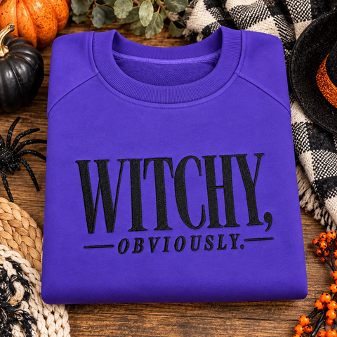 Purple Gildan Witchy Obviously Halloween crewneck sweatshirt with black embroidery displayed with Halloween boutique decorations.
