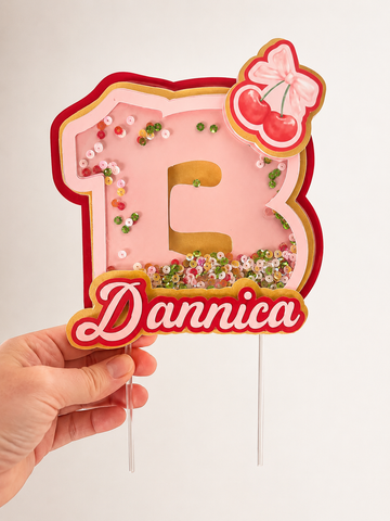 Personalized cherry bow 3D shaker cake topper featuring custom name and age with layered cardstock design and acrylic cake stakes for birthday celebrations.